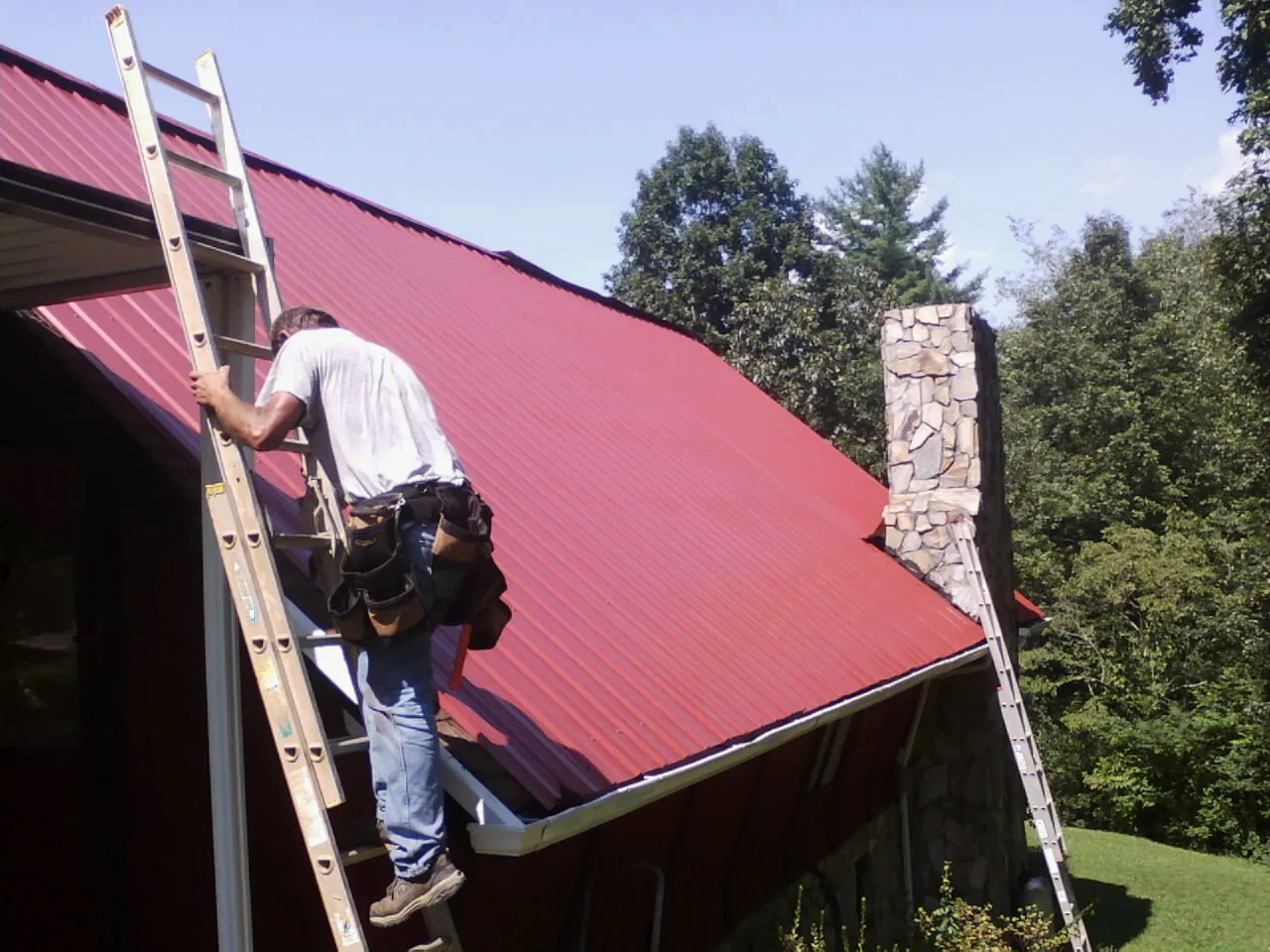 Roofer on a ladder beside a red metal roof and stone chimney — Soffit & Fascia Repair by Roofing Brothers in Fort Blackmore